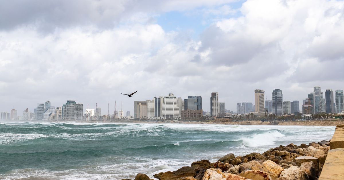 Scenic view of Tel Aviv's skyline with rocky seashore and waves crashing, under a cloudy sky.